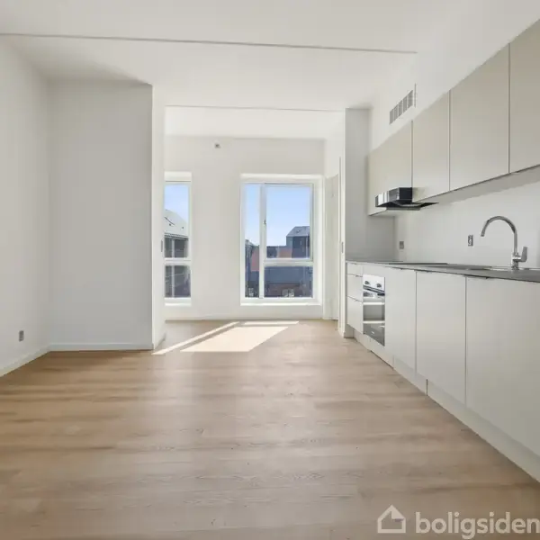 Køkken med hvide skabe og indbygget ovn, står i en åben, lys stue med trægulve. Store vinduer i baggrunden lader naturligt lys strømme ind.