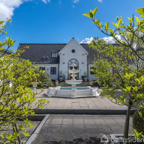 En hvid villa med et centralt springvand står omgivet af grønne buske, under en klar blå himmel.