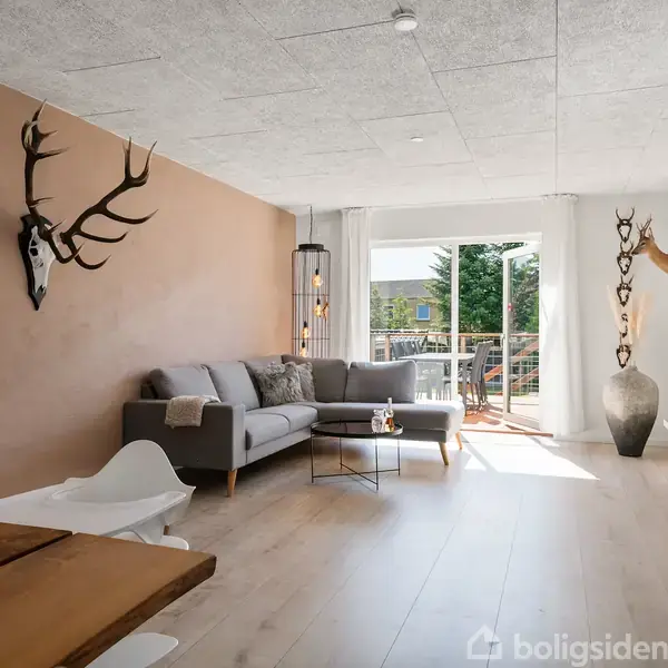 Stue med grå sofa og kaffebord ved en åbning til en balkon. Et hjortegevir pryder væggen, og et såkaldt jordklode står på et skab. Ved siden af sofaen er et stort gulvvase placeret foran en lysebrun væg.