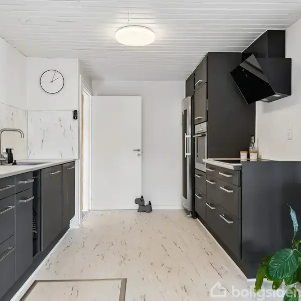 Et moderne køkken med sorte skabslåger og en hvid bordplade. En vask med armatur er monteret, og der hænger et ur på væggen. En grøn plante står i hjørnet.