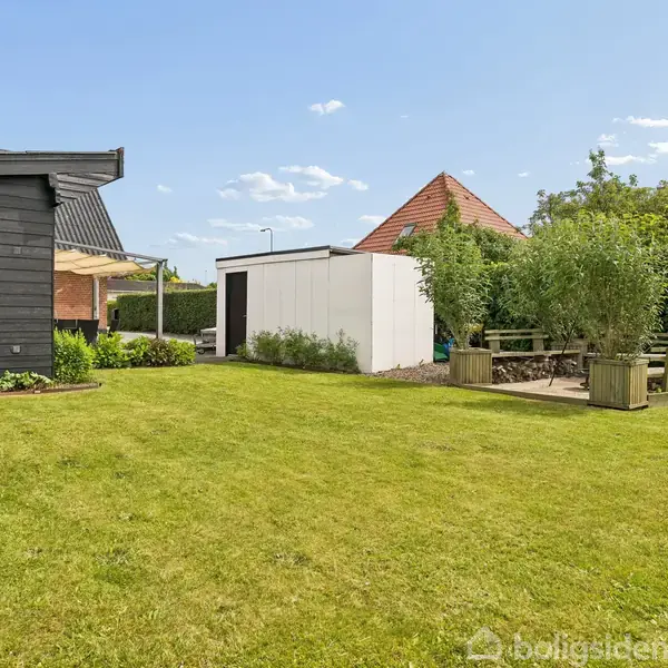 En velplejet græsplæne sidder foran et sortmalet hus. Ved siden af er et hvidt skur; der er et picnicområde omgivet af grønne hække og træer under en klar blå himmel.