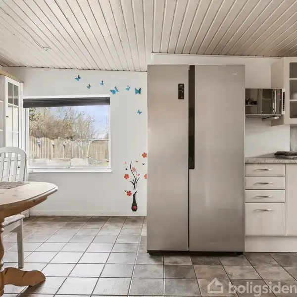 Køleskab står stille i lyst køkken, omgivet af hvide skabe og spisebord med stole. Blå sommerfugle dekorerer væggen ved siden af vindue med udsigt til have.