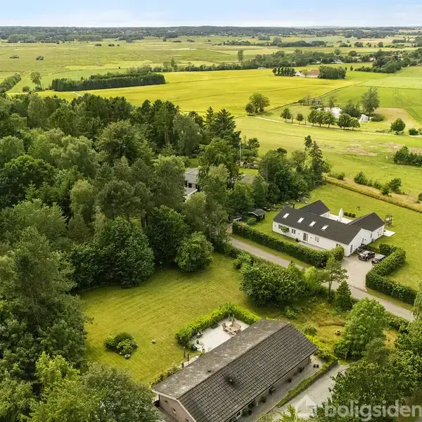 Landområde med et hvidt hus og en lade omgivet af tæt skov og marker. Veje snor sig gennem landskabet, og horisonten viser brede, åbne marker under en klar himmel.