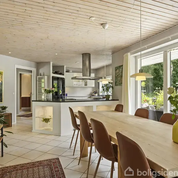 Et træbord med otte stole står i et moderne køkken-alrum. Store vinduer giver udsigt til en grøn have. Kunstværker pryder væggene, og en vase med blomster står på bordet.