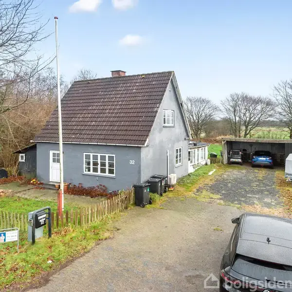 Et gråt hus med skråt tag står i en gård omgivet af græsplæne og træer. Der er tre biler parkeret, og en flagstang står ved siden af huset.
