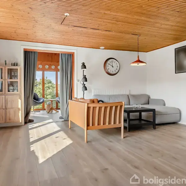 Stue med gråt sofaarrangement og sofabord, vægur på væggen, lamper, reol og vitrineskab. Vindue og dør til veranda med udsigt til grønne træer. Trægulv og loft.