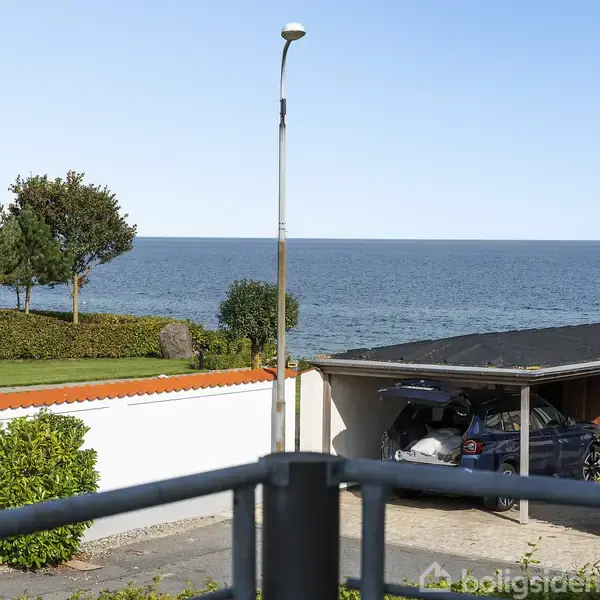 Bil holder i carport; ved siden af en hvid mur med rødt tag. Foran ses græsplæne med træer og buske. I baggrunden er der et klart, blåt hav og himmel.