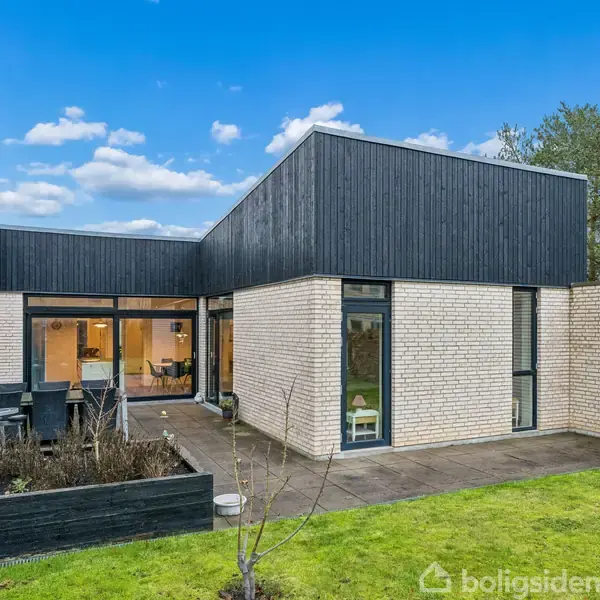 Et moderne hus med lyse mursten og fladt tag. Huset har store vinduer og en terrasse, omgivet af en grøn have med buske og træer under en blå himmel.
