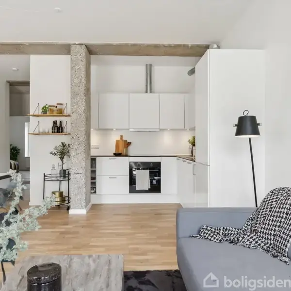 En moderne, lys stue med et åbent køkken. Sofagruppen har en grå sofa og dekorative puder. Køkkenet har hvide skabe og stålapparater. Indretningen er minimalistisk med et lille spisebord.