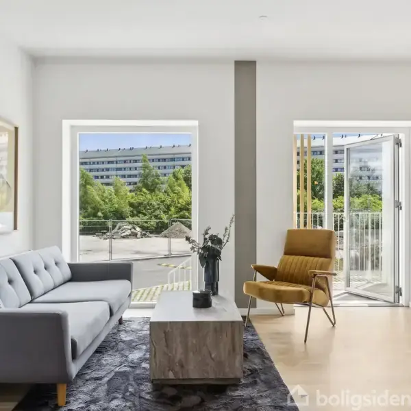 Sofa og lænestol står i en lys stue med trægulv og moderne indretning. Store vinduer og en glasdør åbner ud til udsigt over grønne træer og en bygning udenfor.