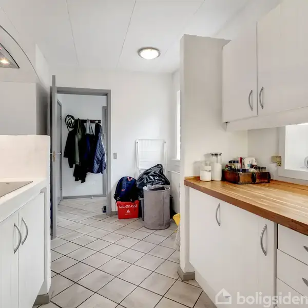 Køkkenet har hvide skabe og en træbordplade. En håndvask er indbygget i bordet. I baggrunden ses en stumtjener med tøj. Flisegulv og en loftslampe oplyser rummet.