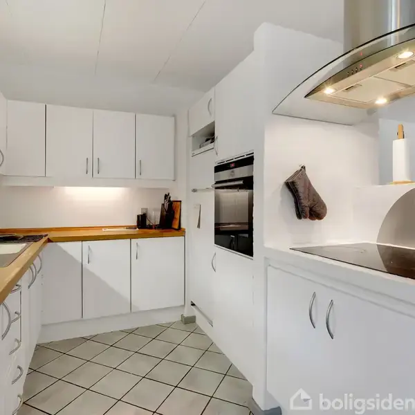 Et køkken med hvide skabe og skuffer. Induktionskogeplade og emhætte er synlige. Bageplade med viskestykke hænger ved ovnen. Flisegulv og bordplader i træ. Vaskemiddel ved vasken.