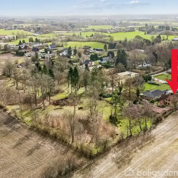 Rødt hus ses fra luften med en stor rød pil pegende på det. Det er omgivet af træer og marker i et landligt område med andre spredte bygninger.