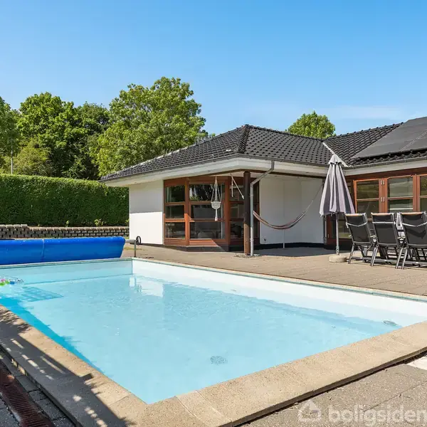 Swimmingpool med klart vand ligger foran et moderne hus med sort tag. Have med tætklippet hæk omkranser pool-området. Havemøbler og parasol står på terrassen ved siden af poolen.