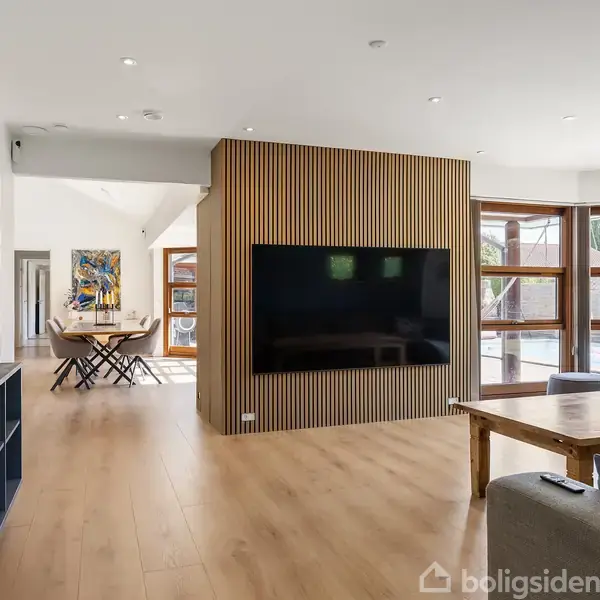Et moderne stueindretning med en stor fladskærm på en trækernevæg. Et spisebord med stole ses i baggrunden ved siden af store vinduer, der åbner mod en terrasse.