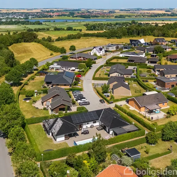 Boligkvarter med moderne huse har en snoet vej i et frodigt landskab. Huse med haver er omgivet af marker og skove i baggrunden.