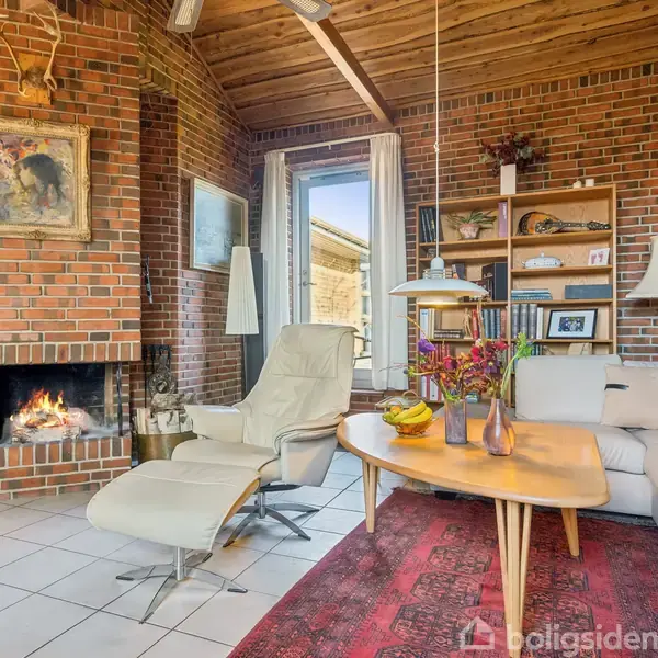 En entre har murstensvægge, et indgangsparti med glasdør og flisegulv. En kommode står ved siden af en knagerække med jakker. En vase med grene står på bordet.