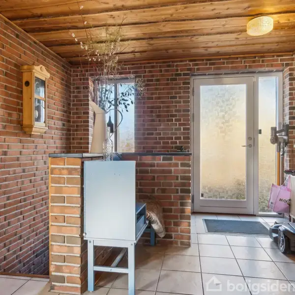 Pejs med ild brænder i hjørne af stue med murstensvægge, lænestol og sofabord med frugtskål og blomster. Rummets loft er træbeklædt, og der er røde tæpper på gulvet.