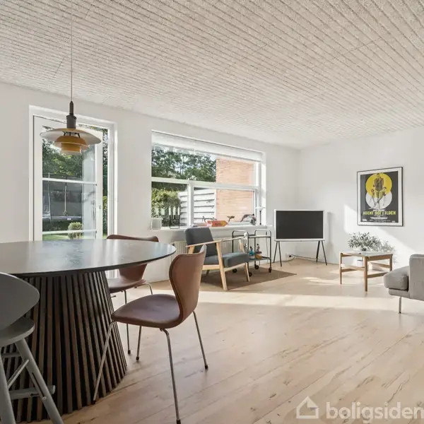 Rundt spisebord med fire stole står i en lys stue. Sollys strømmer gennem store vinduer. Sofa, fjernsyn og farverig vægkunst giver en hyggelig atmosfære.