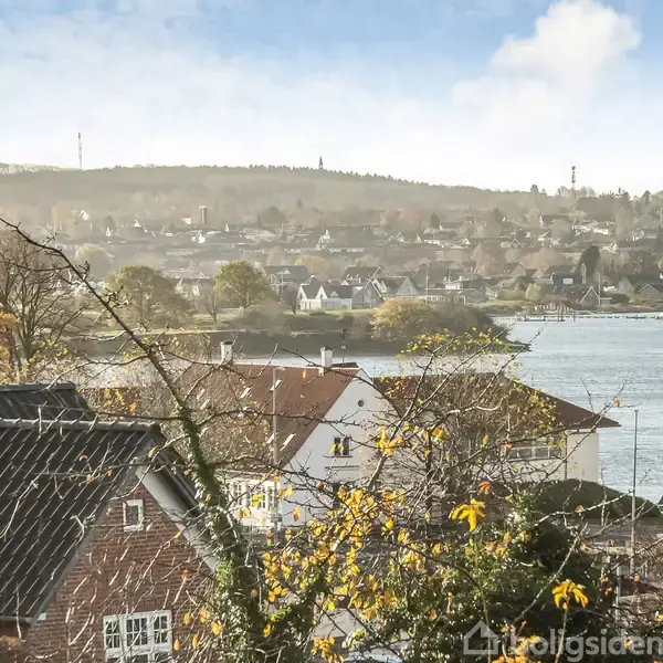 Huse står stille med tage dækket af vægge, omgivet af træer med gule blade. I baggrunden ses en flod og en by ved horisonten under en blå, let overskyet himmel.