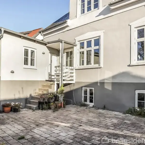 Huset har grå og hvide facader med flere vinduer. Trappe fører til en lille terrasse. Omgivet af fliser, planter og grønne klatreplanter på en mur.