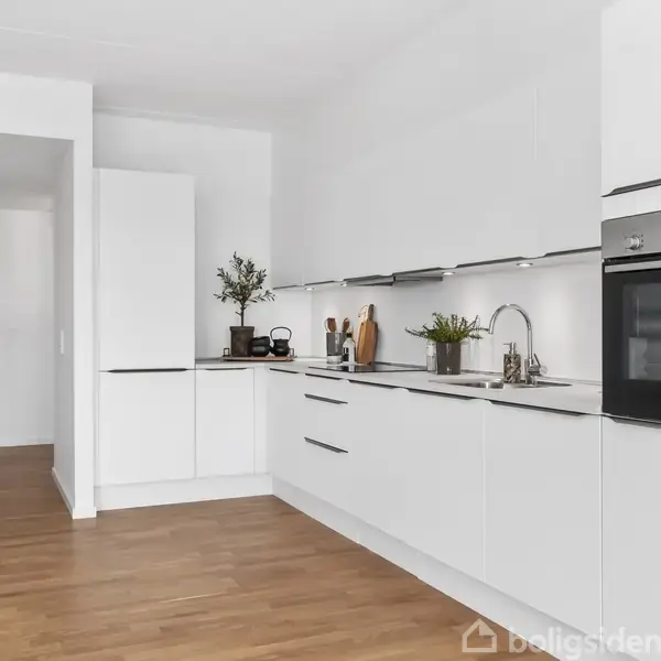 Et moderne, hvidt køkken med indbyggede skabe, ovn og vask. Køkkenredskaber og planter dekorerer bordpladen. Rummet har trægulv og hvidmalede vægge, hvilket skaber en lys og ren atmosfære.