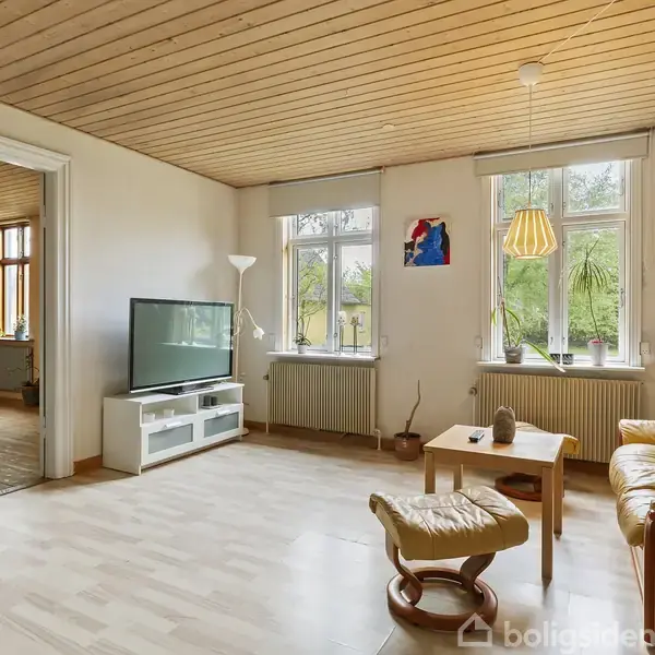 Stue med en beige lædersofa og fodskammel, et lille træbord og et tv på hvidt møbel. Store vinduer bringer naturligt lys ind i det lyse rum.