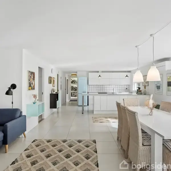 En moderne stue med et blåt hjørnesofa, der står ved siden af et farverigt maleri. I baggrunden er et spisebord med lysebrune stole og hængende lamper.