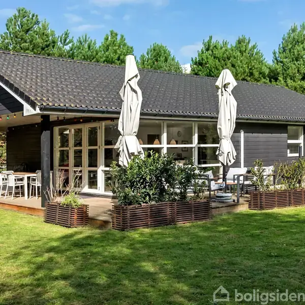 Et sommerhus med sorte trævægge og terrasse, møbleret med borde og stole. Paraplyer står sammenfoldet. Omgives af grønne græsplæner og tætbevokset skov i baggrunden.