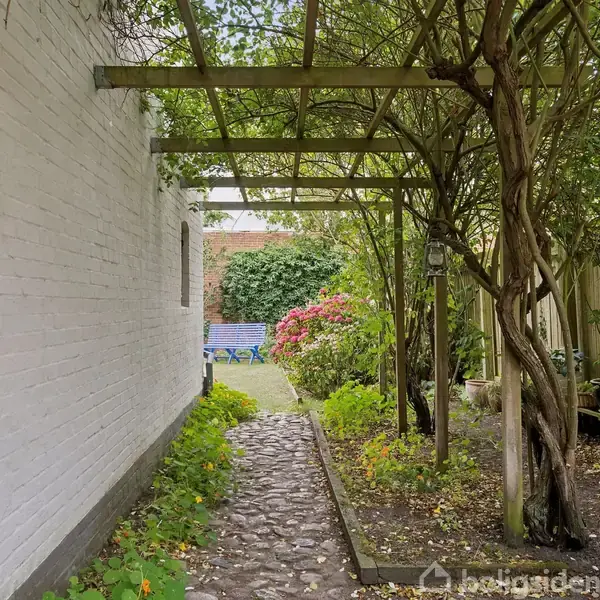 Stenbelagt sti strækker sig gennem haven, dækket af trælpergola med klatrende planter. Til højre ses buske og blomster, mens en blå bænk står for enden.