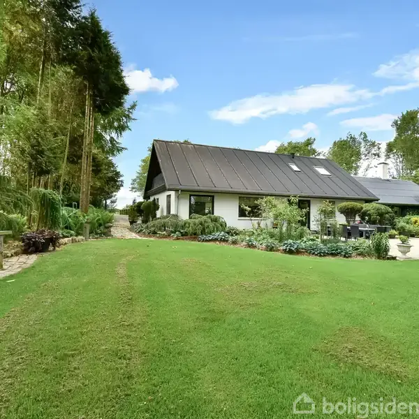 Et hus med sort tag står omgivet af en velplejet have med grøn græsplæne og træer i baggrunden. Ved siden af huset er blomster og buske plantet langs husets facade.