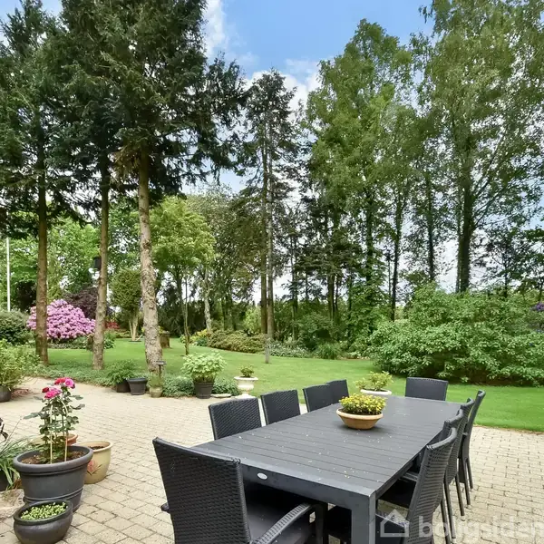 Spisebord i sort udendørsmateriale står på en terrasse omringet af blomsterkrukker og grønne planter. Haven i baggrunden har græsplæne og høje træer under en blå himmel.