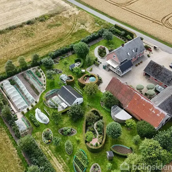 En gård med flere bygninger og et omfattende haveanlæg. Bygningerne ligger ved en landevej, omgivet af marker. Haven har drivhuse, bænke og varieret beplantning, omkranset af træer og buske.