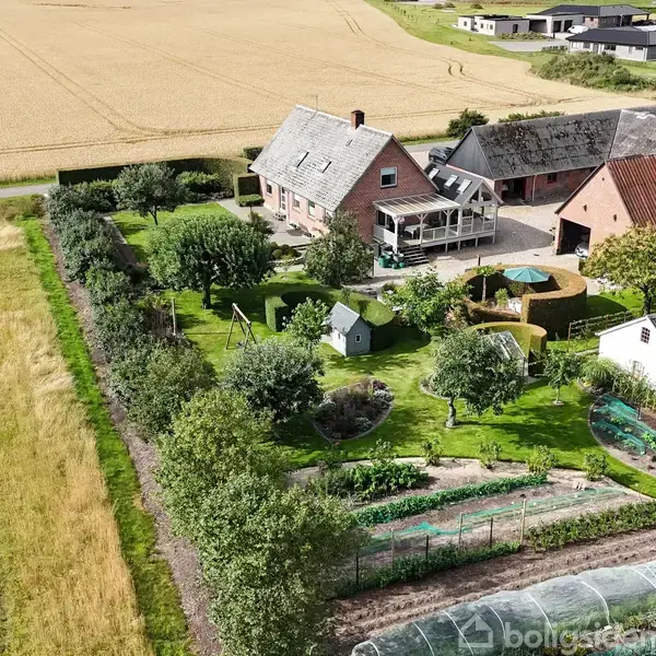 Huset er omgivet af en frodig have med frugttræer og små haver. Det ligger i landlige omgivelser med marker omkring. Adskillige udhuse er synlige ved siden af hovedbygningen.