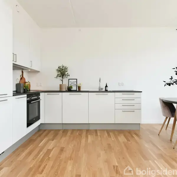 Et hvidt, moderne køkken med indbyggede skabe og sort bordplade. Ovn og vask er installeret. Spisebord med stole står til højre, dekoreret med planter. Trægulv.