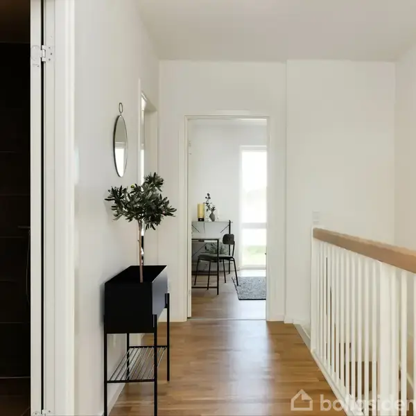 Gang med trægulv, et sort konsolbord med plante og rundt spejl. Dør til højre fører til lyst rum med bord og stole. Ingen tekst fundet.