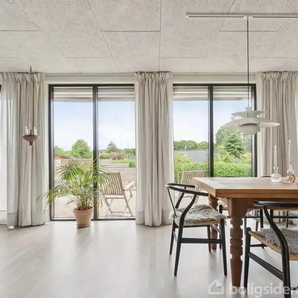 En spisestue med et træbord og stole, omgivet af store glasdøre, der åbner ud til en terrasse med planter. Lyse gardiner hænger fra loftet, og udsigten viser grønne omgivelser.