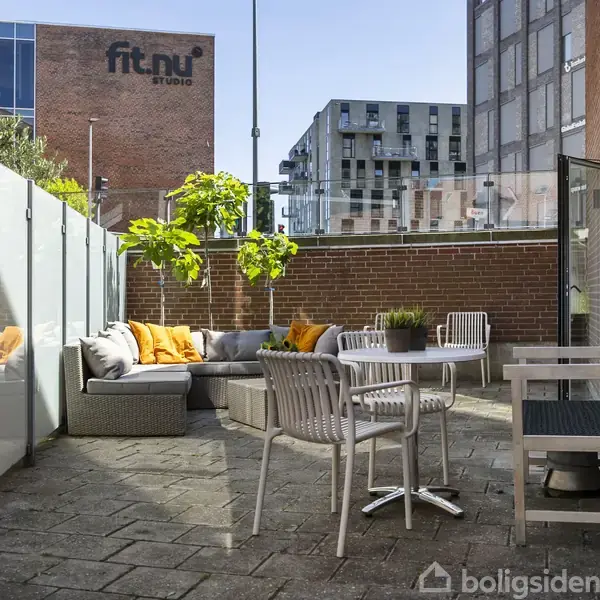 Terrassemøbler står på en flisebelagt altan med glasafskærmning. Borde og stole er omkring sofaen med orange puder. Omkring ses bybygninger, herunder "fit.nu studio" på en murstensbygning.