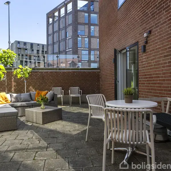 Et møbleret gårdhavemiljø med sofaer, borde og stole. Hallen har murstensvægge, planter og er omkranset af høje bygninger under en blå himmel.