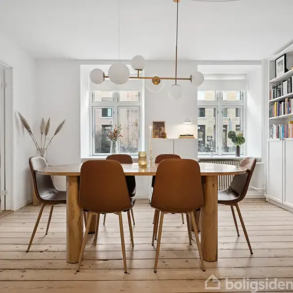 Et spisebord med seks brunlæderstole står på et trægulv. I et lyst rum hænger en moderne lysekrone. Bøger og dekorative genstande fylder hylder på højre væg.