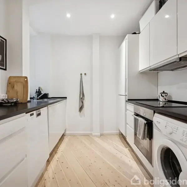 Et moderne køkken med hvide skabe og sorte bordplader, hvor der står en vaskemaskine, mikroovn og andre køkkenredskaber. Trægulv og en viskestykke hænger på væggen.