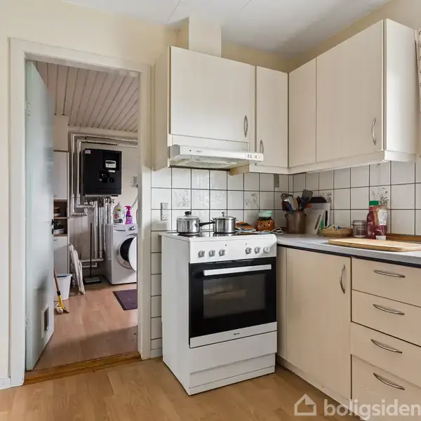 Komfur med gryde på, placeret i et køkken med hvide skabe og flisebelagt backsplash. Til højre ses en lysfyldt vinduesramme med udsigt til en have. Vasketøj i baggrunden.