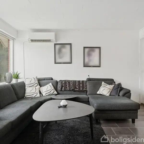 En sort hjørnesofa med hvide puder står i en lys stue. Foran sofaen er et sort sofabord på gulvtæppe. Væggene er hvide med minimalistisk dekoration.