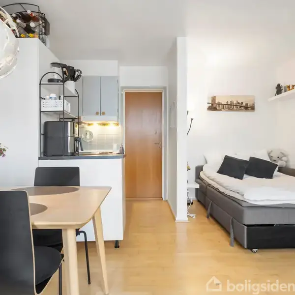 Lejlighed med moderne interiør; spisebord med blomster til venstre, sofa overfor tv til højre. Køkkenhjørne i midten, dør i baggrunden. Hylder med dekorationer på væggen.