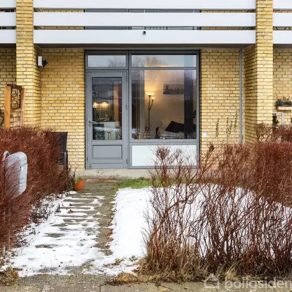 En rækkehusfacade med gule mursten og en stor glasdør foran et lille forhaveområde med få snepletter. Tørre buske flankerer stenstien, og en lanterne står ved siden af døren.