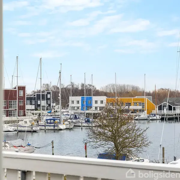 Både ligger stille i en marina omgivet af farverige huse i røde, blå og gule nuancer. En træhytte ses i forgrunden under en klar blå himmel.
