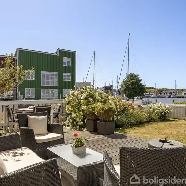 Terrassemøbler står på en trædækket terrasse med udsigt til marinaen. Blomster og planter omringer området, mens en grøn bygning og både ses i baggrunden under en klar, blå himmel.