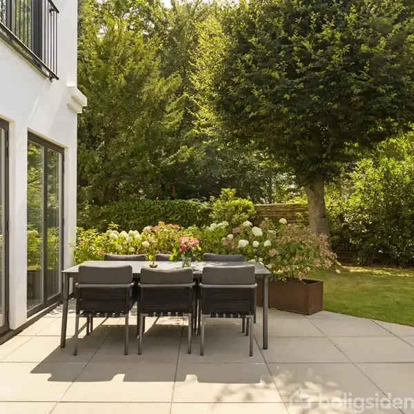 Et havebord med seks stole står på en flisebelagt terrasse ved siden af et moderne hus med glasdøre. Haven er frodig med træer og blomster i baggrunden.