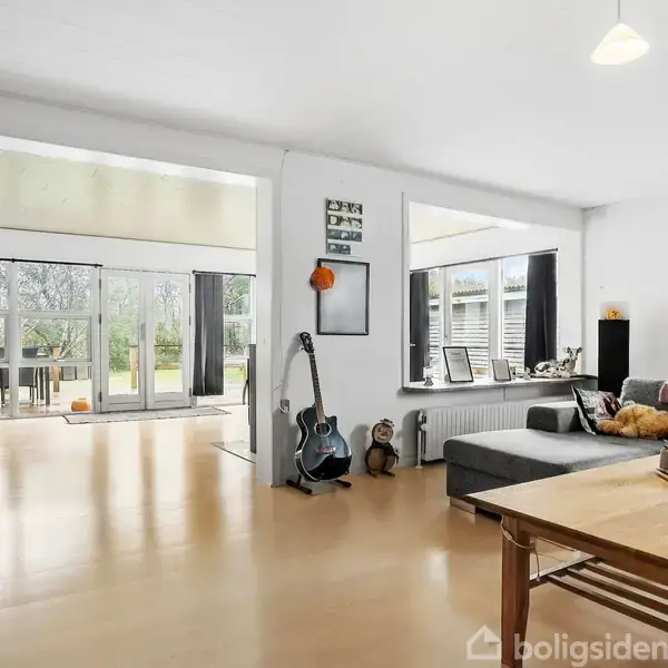 En stue med en grå hjørnesofa står ved en træspisebord. Væggen har kunst og guitarer lent op ved siden af. Store vinduer åbner ud til en terrasse og have.