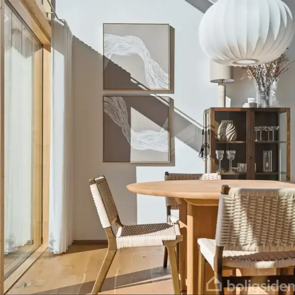 Rundt træbord med fletstole står i solrig spisestue. To abstrakte malerier hænger på væggen. Høj reol med glas og dekorative elementer. Stor hvid loftslampe tilføjer belysning, vinduer til venstre.